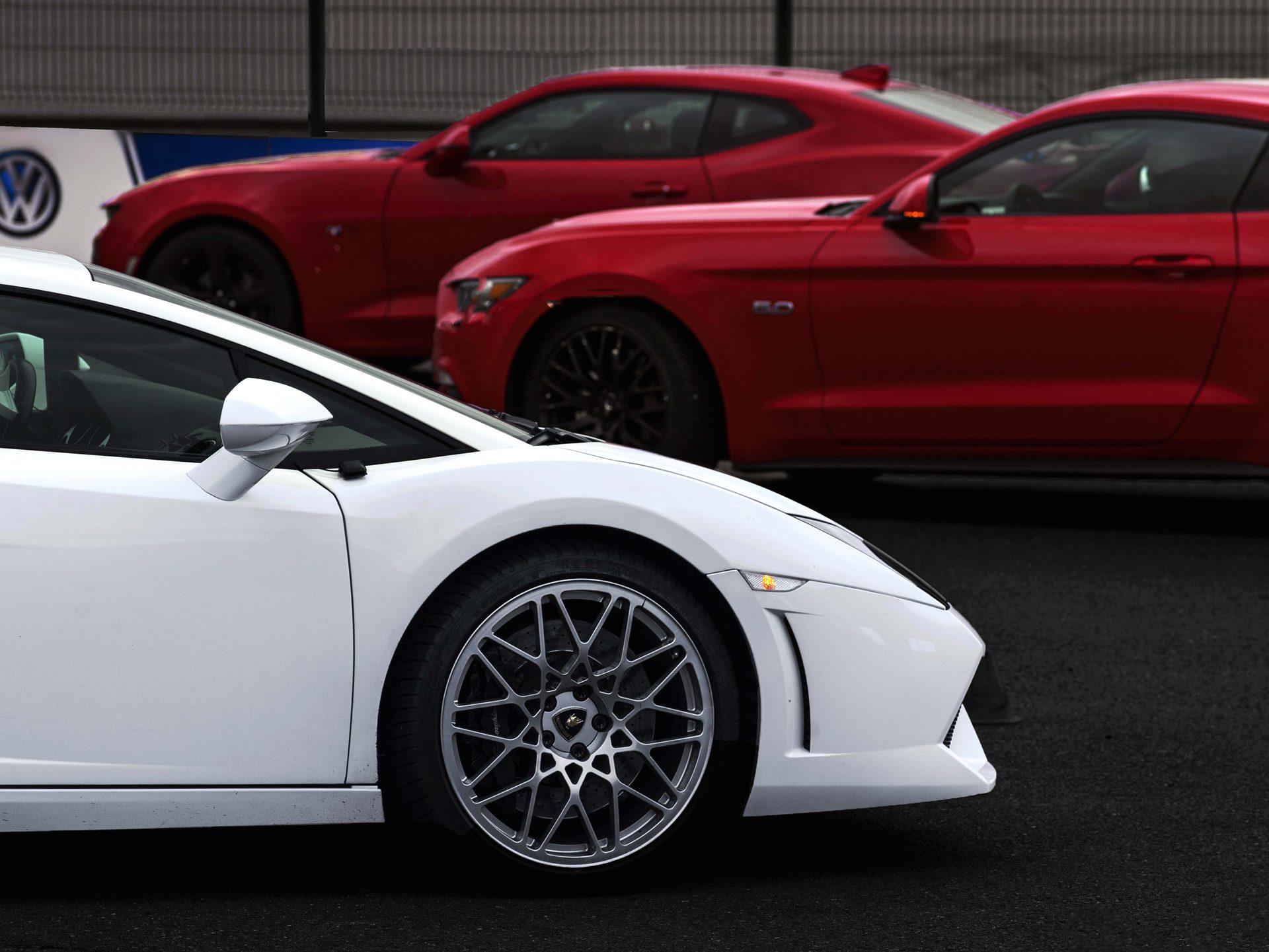 Cascadevents — alignement à l'arrêt : Lamborghini Gallardo blanche au premier plan et Ford Mustang rouges en arrière-plan, parking du circuit