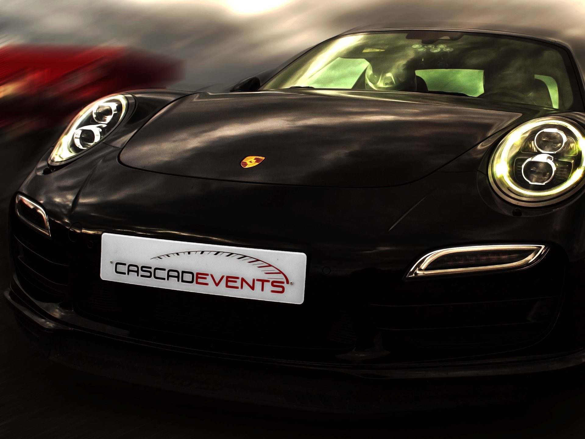 Porsche 911 Turbo noire en gros plan trois quarts avant, optique LED ronde diurne allumée, ciel travaillé en arrière-plan