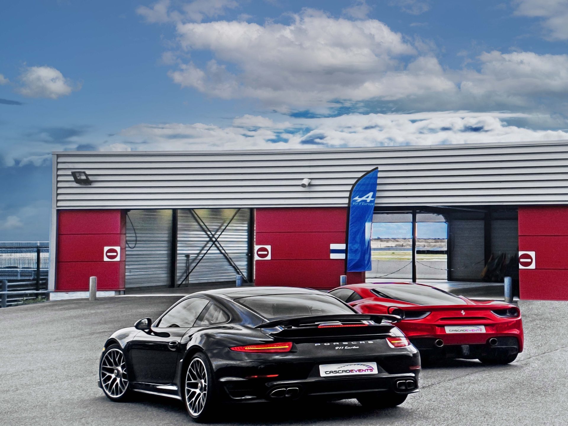 Porsche 911 Turbo noire et Ferrari 488 rouge côte à côte devant les hangars rouges du circuit de La Ferté-Gaucher, ciel bleu nuageux travaillé