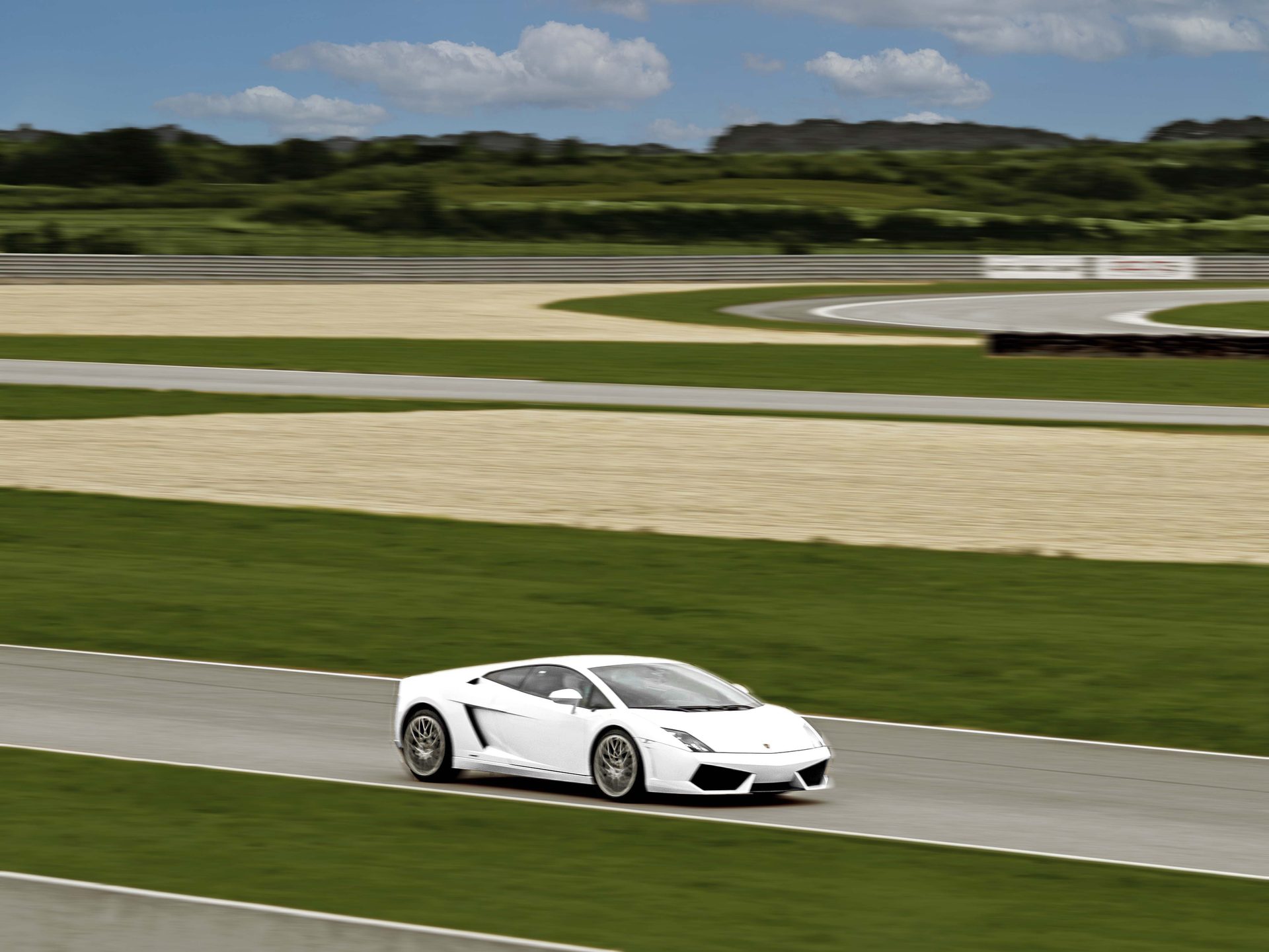 Après retouche : ciel bleu nuagé, végétation vert vif, lumière chaude sur la carrosserie de la Lamborghini Gallardo blanche en filé sur la piste
