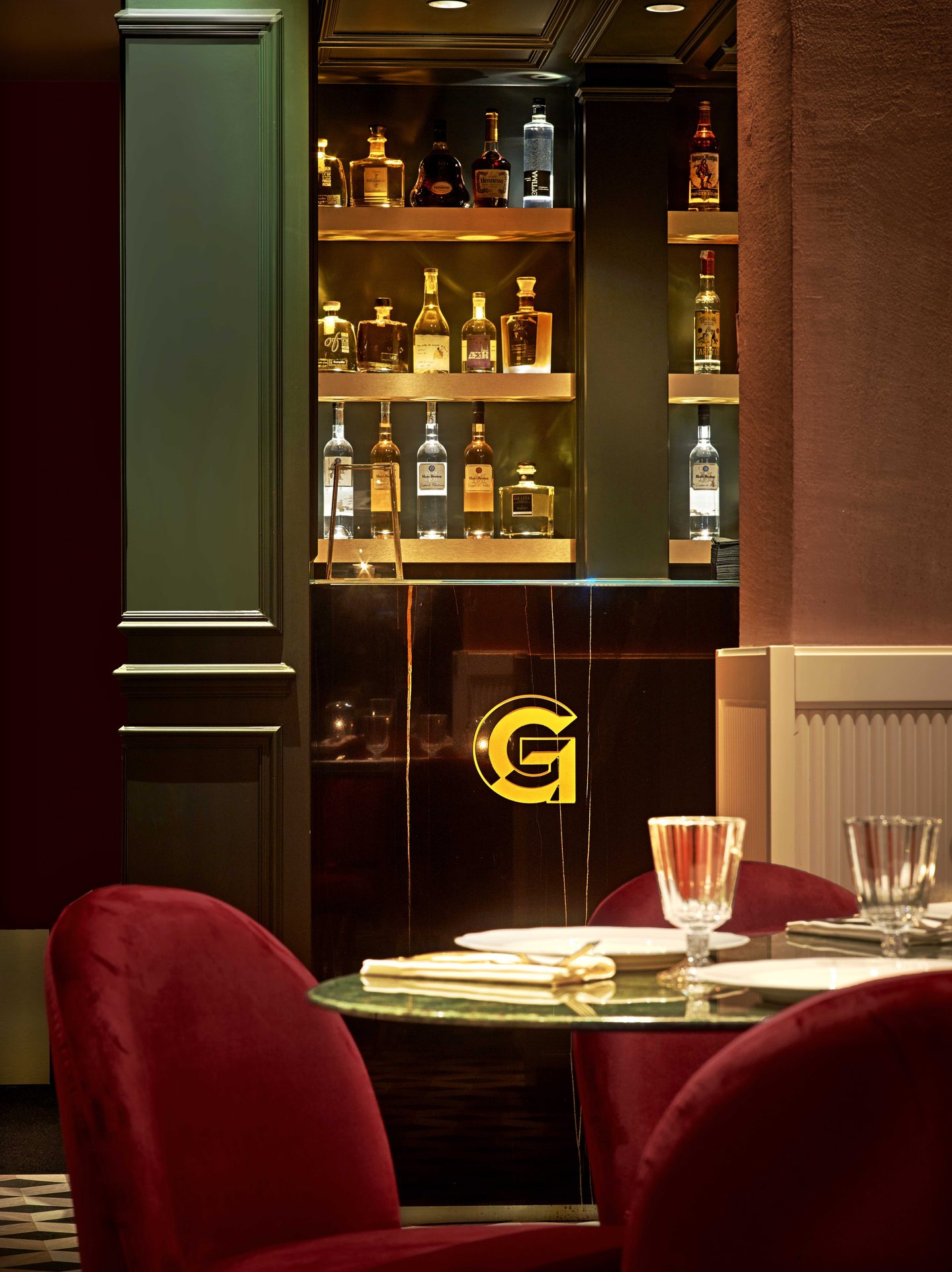 Détail du bar G : logo doré sur laque noire, étagères de bouteilles éclairées en or, chaises velours rouge au premier plan