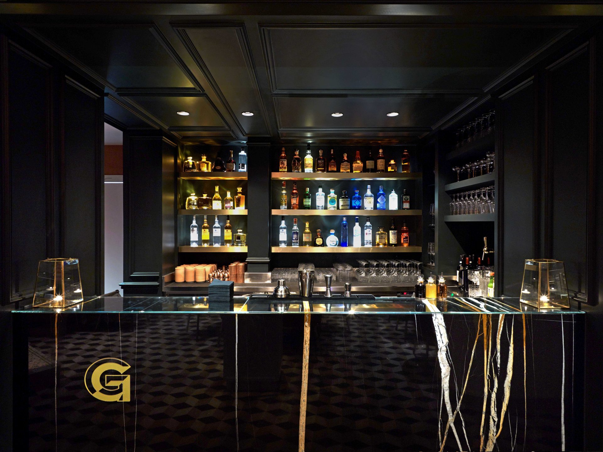 Bar G frontal : comptoir marbre noir veiné d'or, deux lanternes en laiton aux extrémités et bouteilles alignées en arrière-plan
