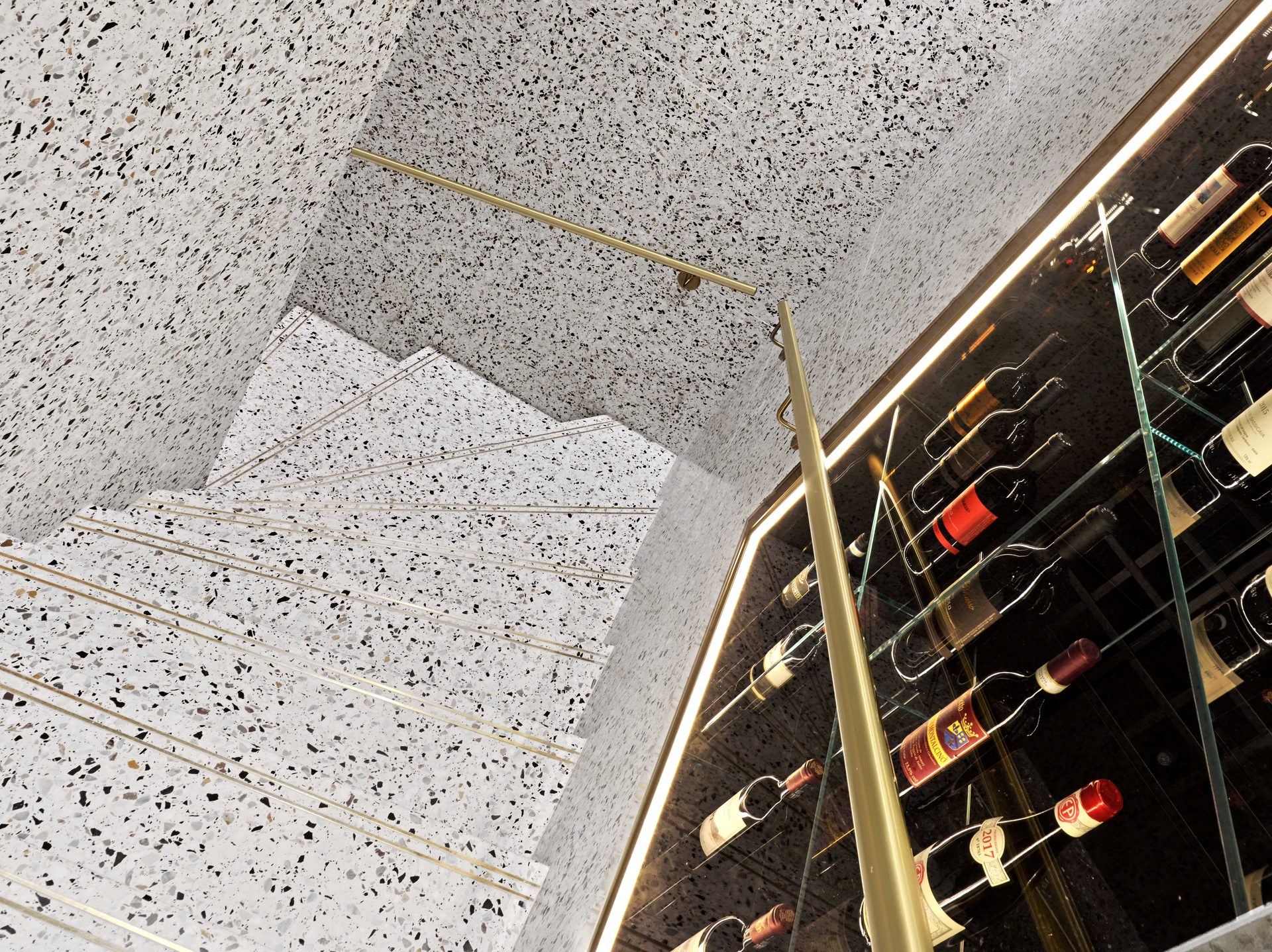 Cave à vins terrazzo vue en contre-plongée, rambarde laiton et bouteilles italiennes éclairées