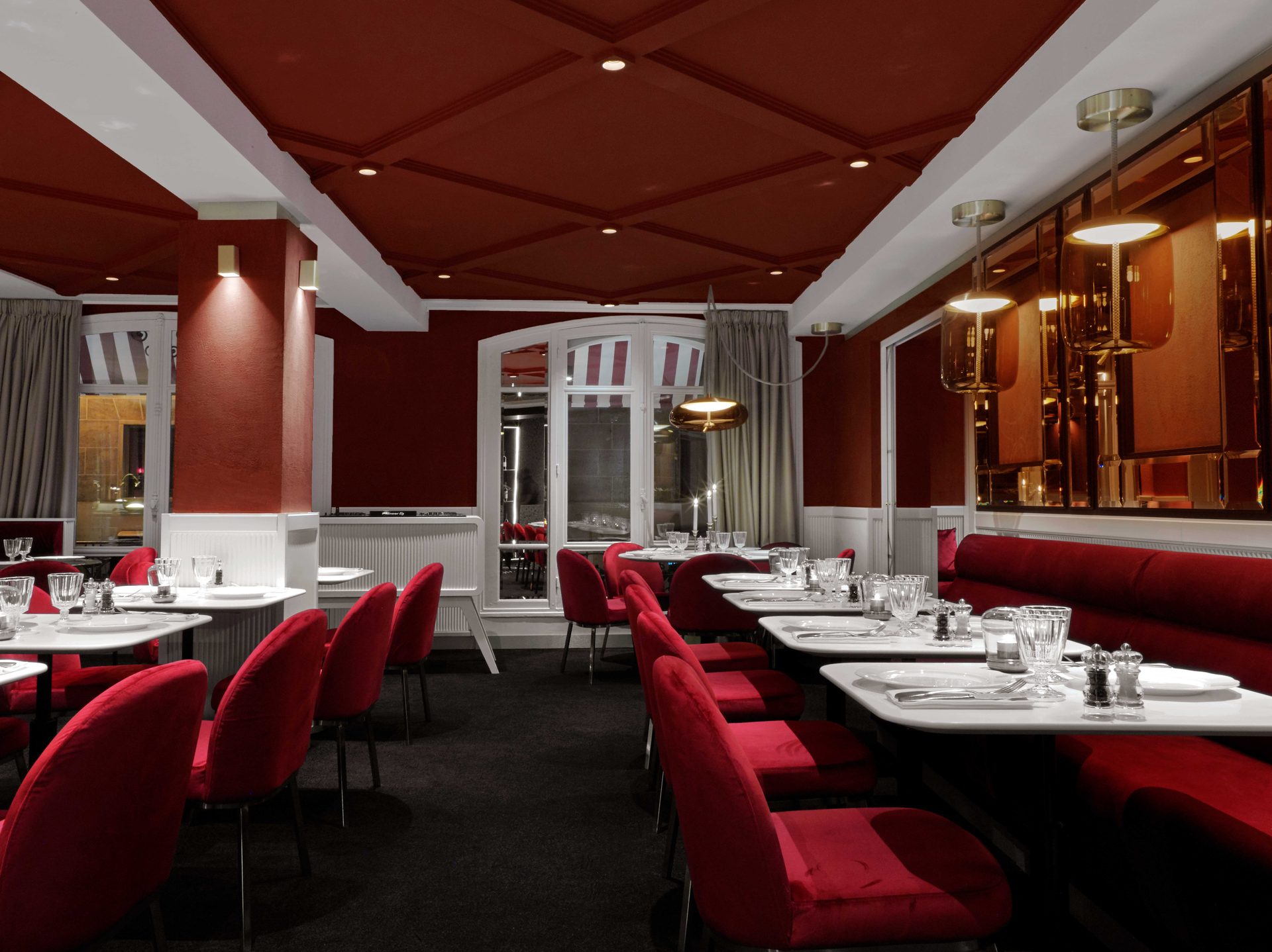 Salle rouge dressée : tables blanches, banquette en velours, suspensions ambre et candélabres allumés