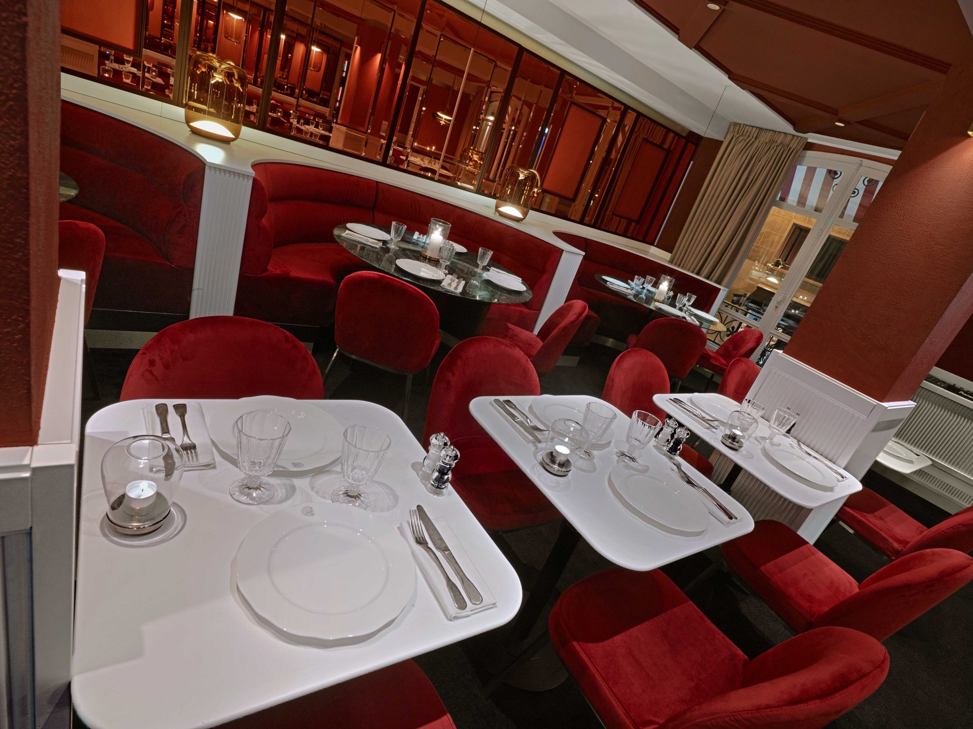 Salle rouge haute en plan large : banquettes rouges, tables blanches dressées et bandeau de miroirs cuivrés