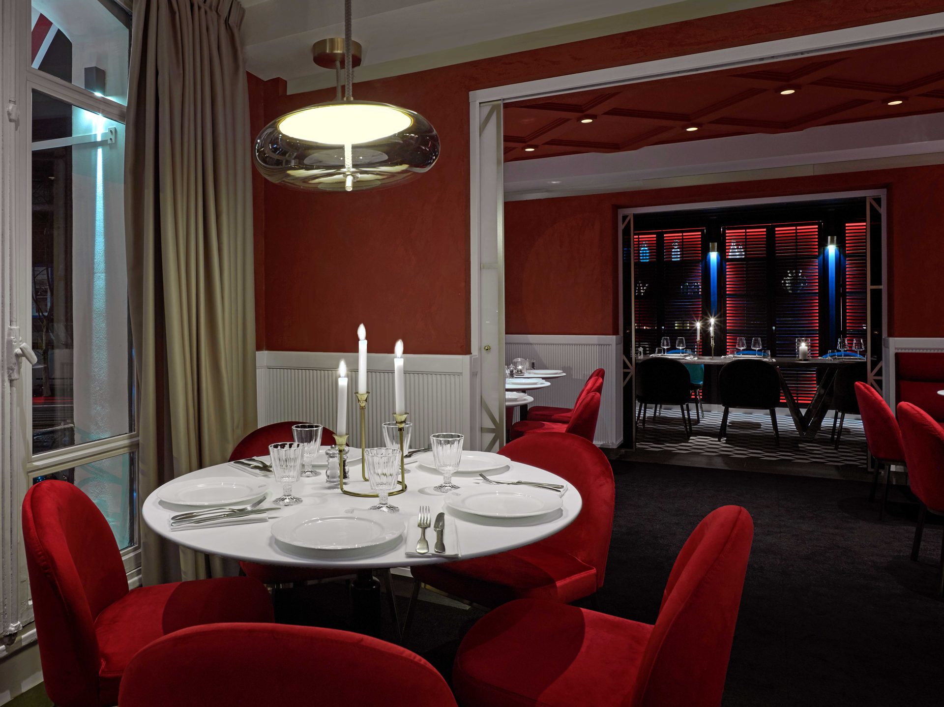 Salle haute rouge avec table ronde au candélabre allumé, ouverture en enfilade sur le privatif noir aux stores rouges rétroéclairés