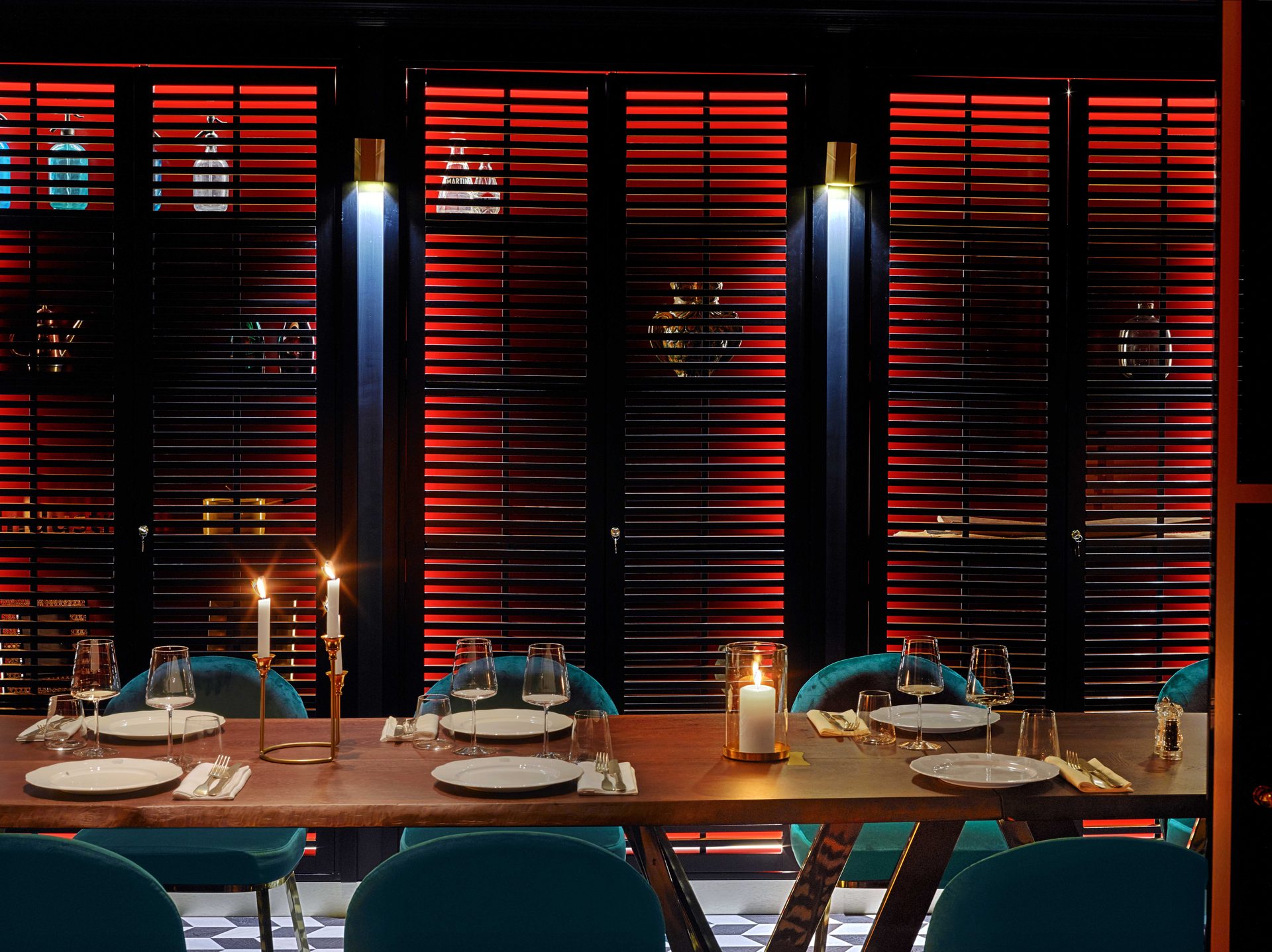 Privatif noir vu frontalement : longue table de bois brut, chaises velours bleu canard, candélabres allumés et stores à lamelles rouges rétroéclairés