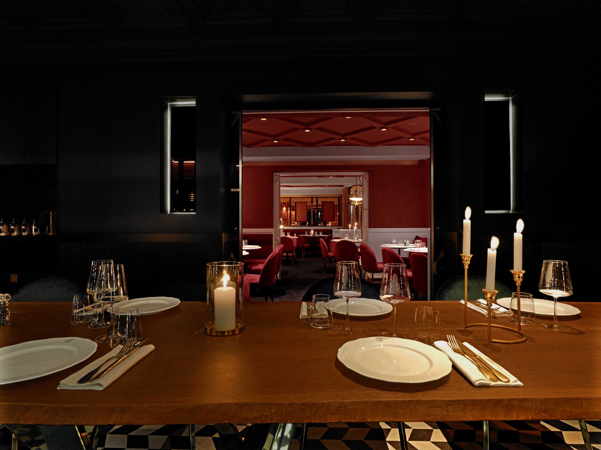 Guisé Trattoria — séquence privatif noir 1/3, ouverture frontale sur la salle rouge en arrière-plan, table de bois brut et candélabres allumés au premier plan