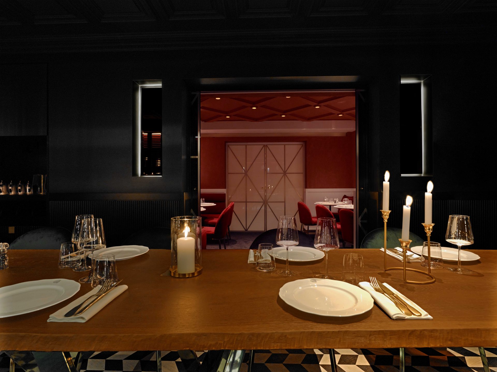 Guisé Trattoria — séquence privatif noir 2/3, porte à motif étoilé en laiton fermée au fond, table de bois brut et candélabres allumés au premier plan