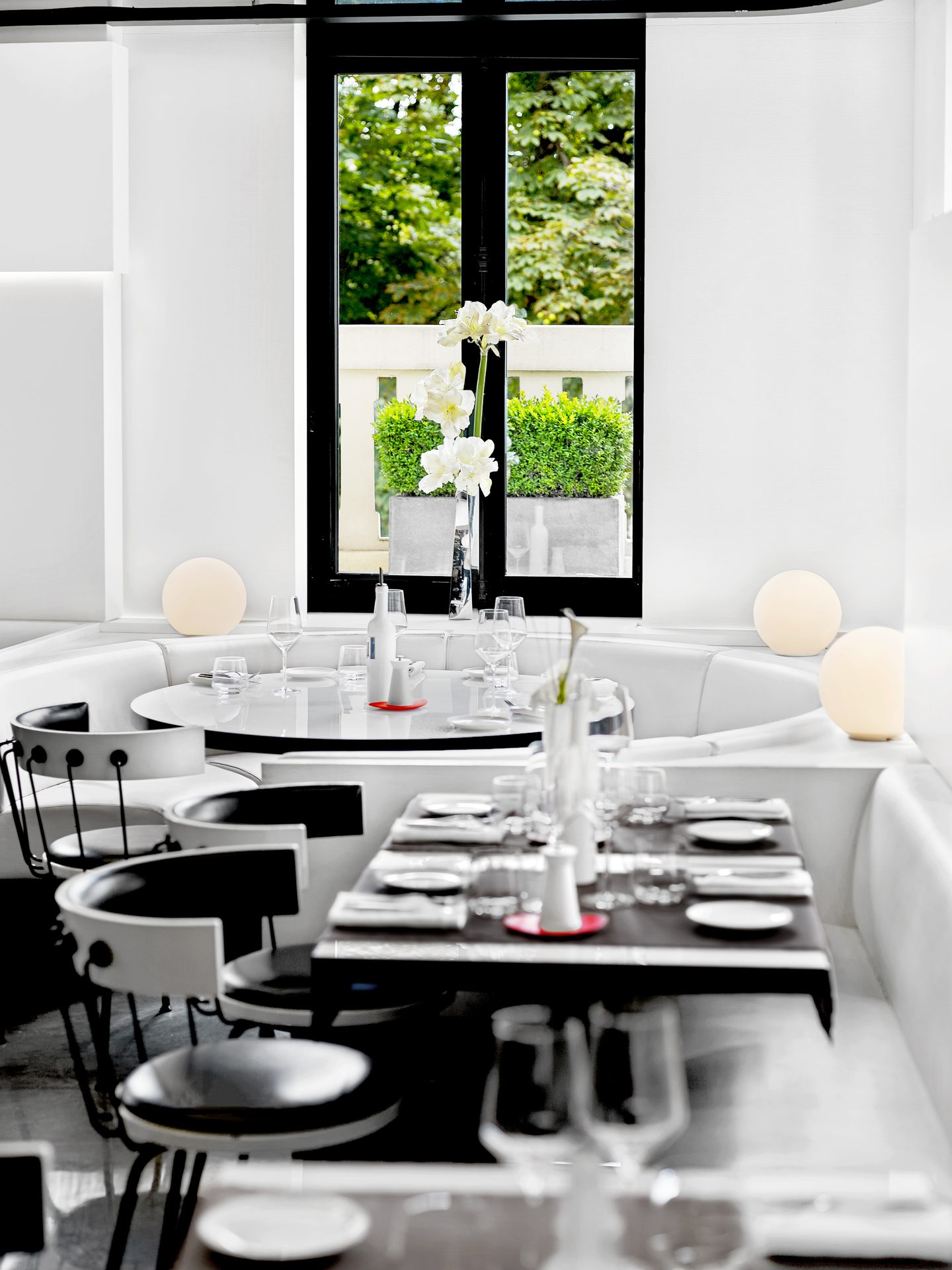 Nolita Paris — vue 04 — cadrage vertical sur la table ronde en alcôve, fleurs blanches d'amaryllis dans vases inox, chaises blanc et noir, baies vitrées