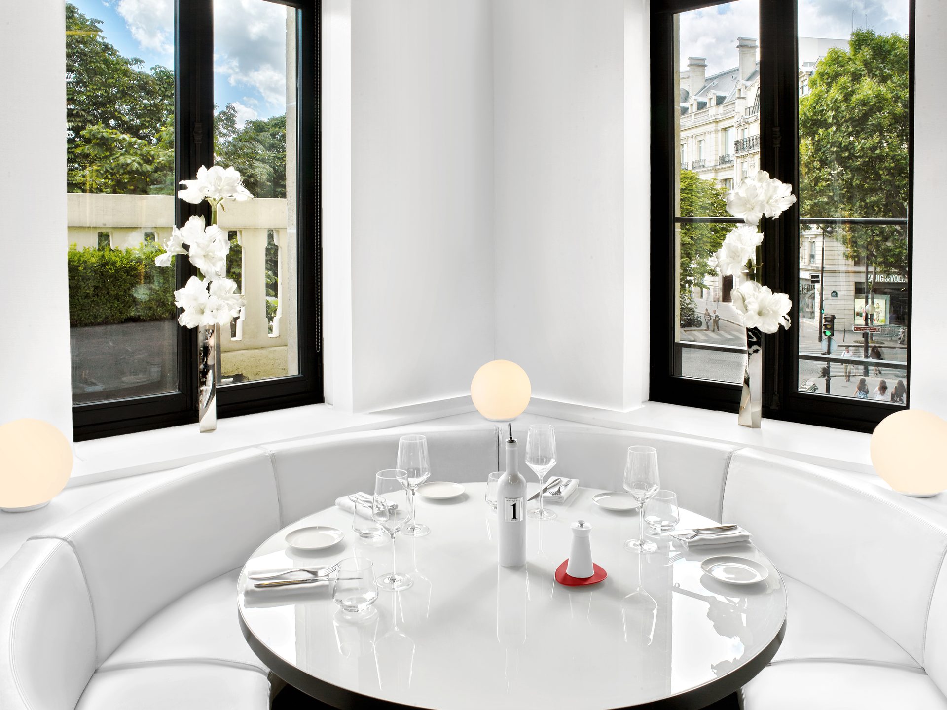 Nolita Paris — vue 06 — alcôve d'angle en bay-window, table ronde blanche pure et banquette circulaire blanche, deux amaryllis aux fenêtres avec vue sur la rue