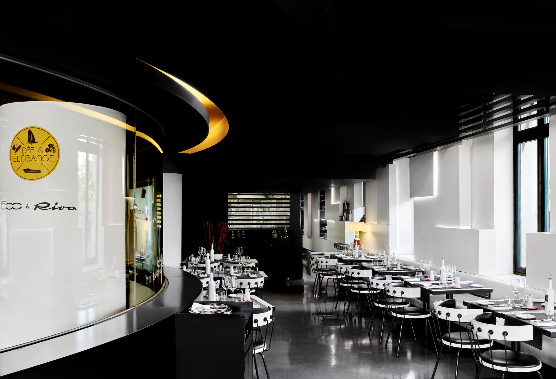 Nolita Paris — vue 18 — alcôve Fiat 500 vue rapprochée, vitre courbe noire au logo 500 & Riva, plancher acajou et calandre Fiat 500C noire, tables blanches cloutées au premier plan