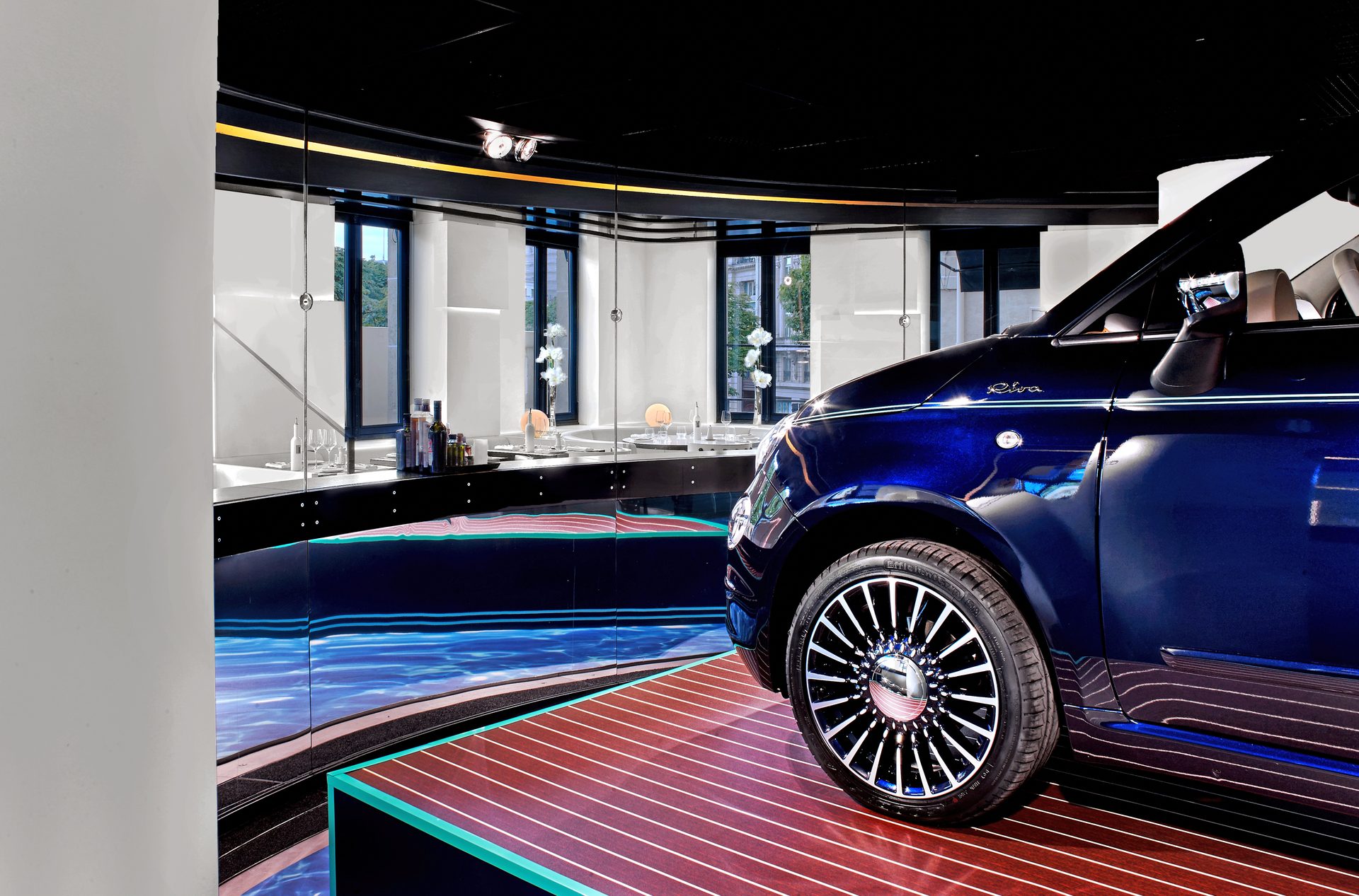 Nolita Paris — vue 34 — alcôve Fiat 500 vue de côté, vitrine noire courbe sur sol effet eau bleue et plancher Riva, miroirs reflétant la Fiat 500 Riva bleu marine