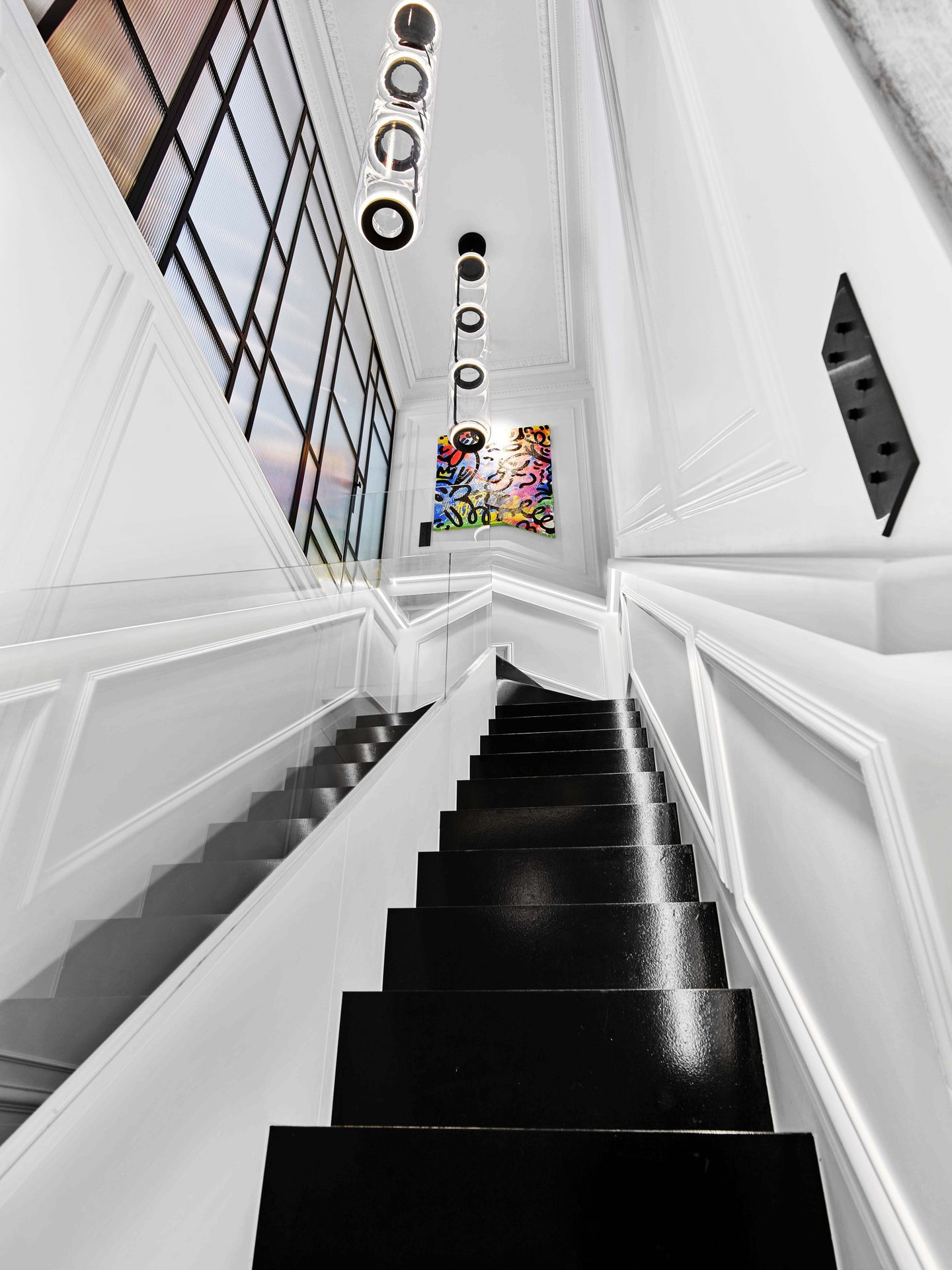 Pereire — vue 09 — escalier laqué noir vu en contre-plongée, verrière, suspensions tubulaires alignées et toile street-art
