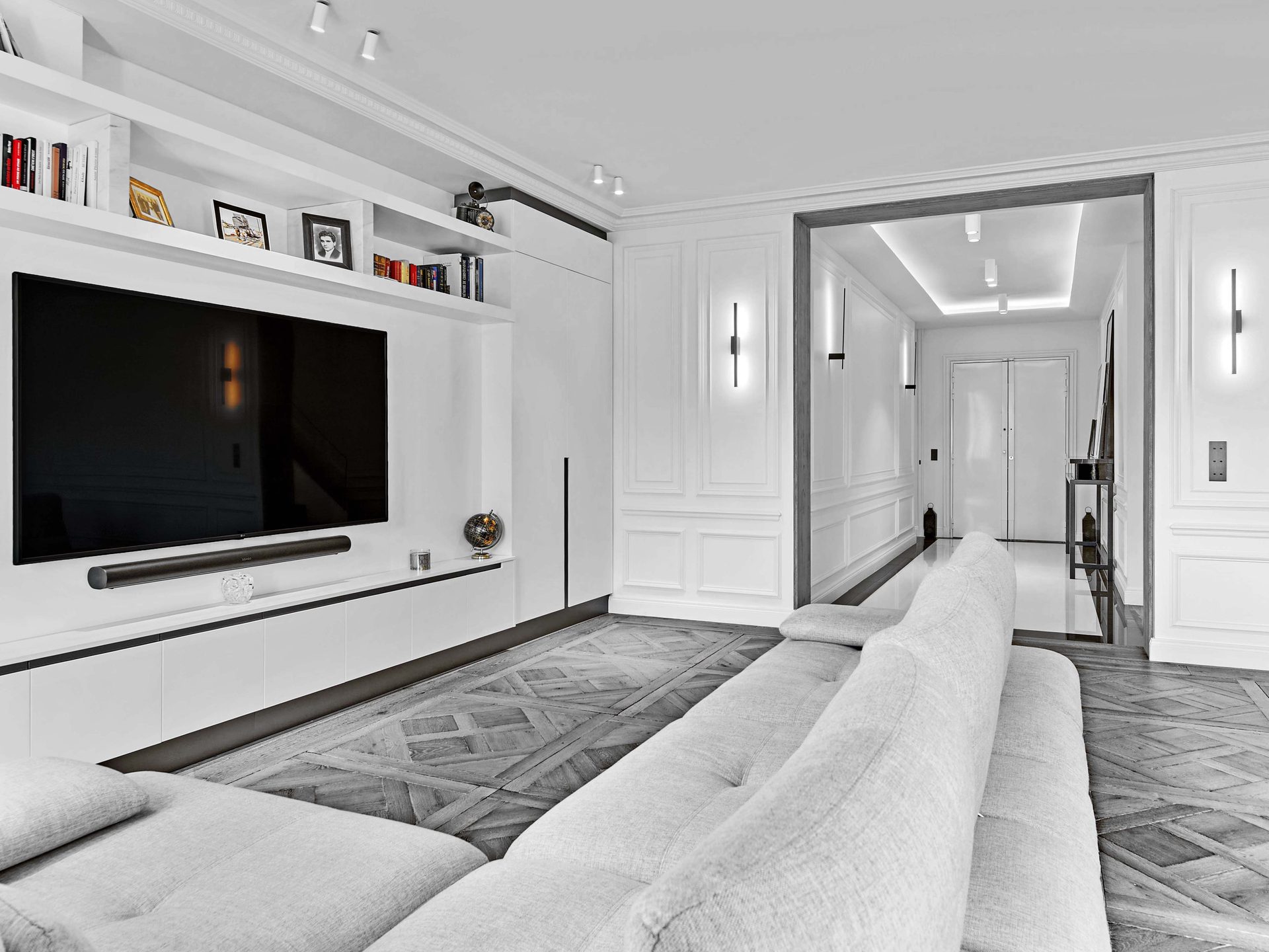 Pereire — vue 21 — séjour, grand canapé d'angle gris, mur TV laqué blanc avec bibliothèque et photographies encadrées, parquet de Versailles, enfilade vers le couloir à boiseries