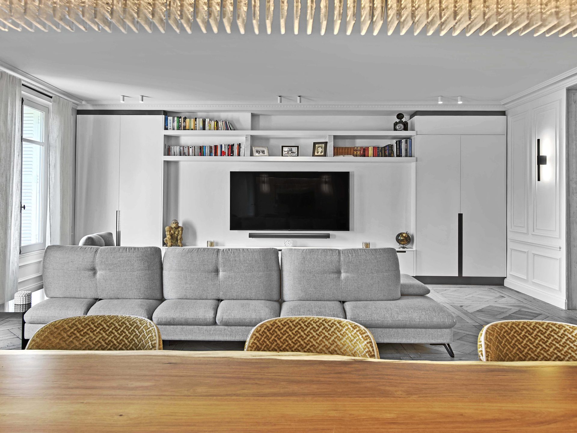 Pereire — vue 24 — séjour vu depuis la table à manger, mur TV laqué blanc et bibliothèque, canapé gris, suspension dorée à baguettes de verre au plafond