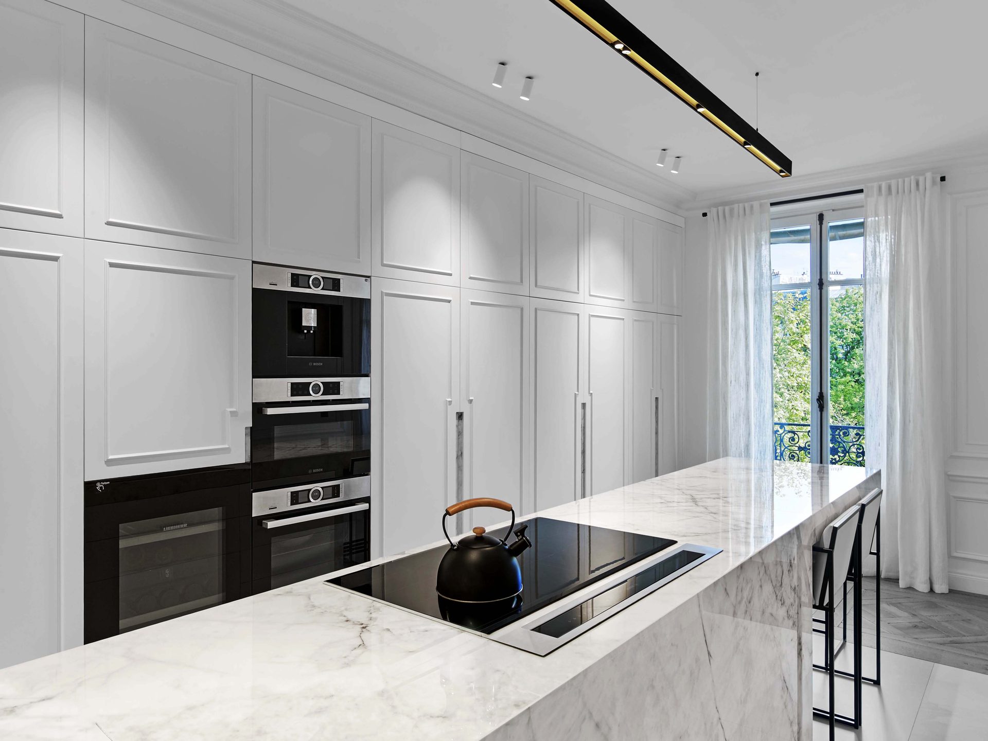 Pereire — vue 41 — cuisine, plan de cuisson sur îlot de marbre, hautes façades laquées blanches et fours Bosch encastrés, fenêtre à voilages sur balcon parisien