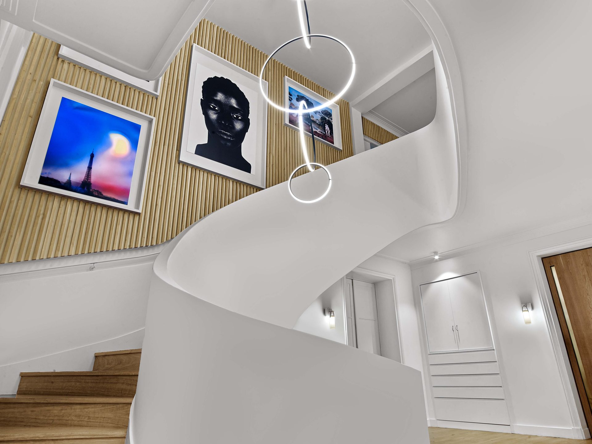 Beaux Arts — vue 08 — escalier blanc sculptural vu en contre-plongée sous les anneaux lumineux, tasseaux de chêne et œuvres encadrées
